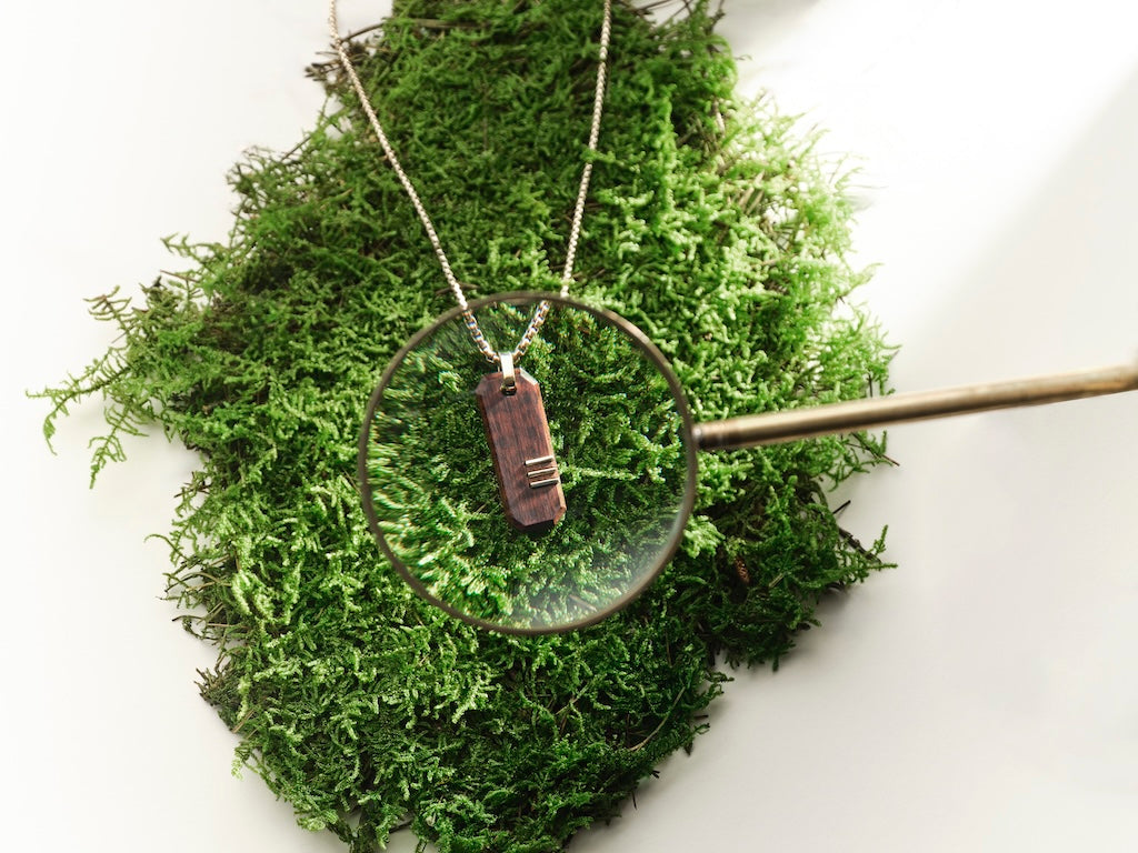 A gold boxchain necklace with a wooden tag displayed on a bed of green moss, with a magnifying glass resting over the pendant and a brass tool extending from the right side, against a white background.