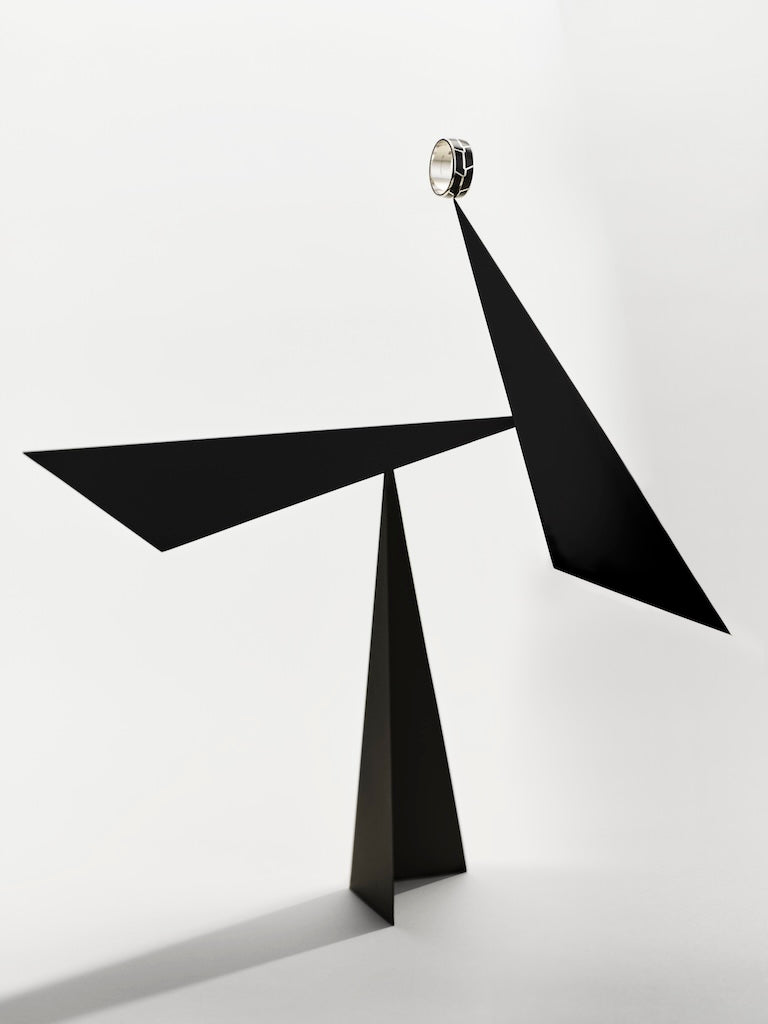 A silver men's band ring with black inlays mounted on top of a minimalist black metal abstract sculpture with sharp geometric triangular forms, mounted against a clean white wall with a small circular metallic accent at the top.