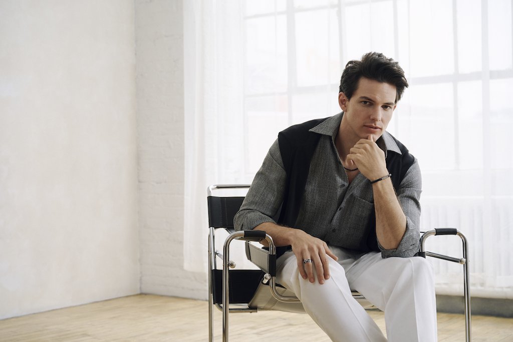 A man wearing a silver and black ring along with a black bracelet and beaded necklace sits in a black and chrome chair in a bright, modern room with white brick walls and large windows.