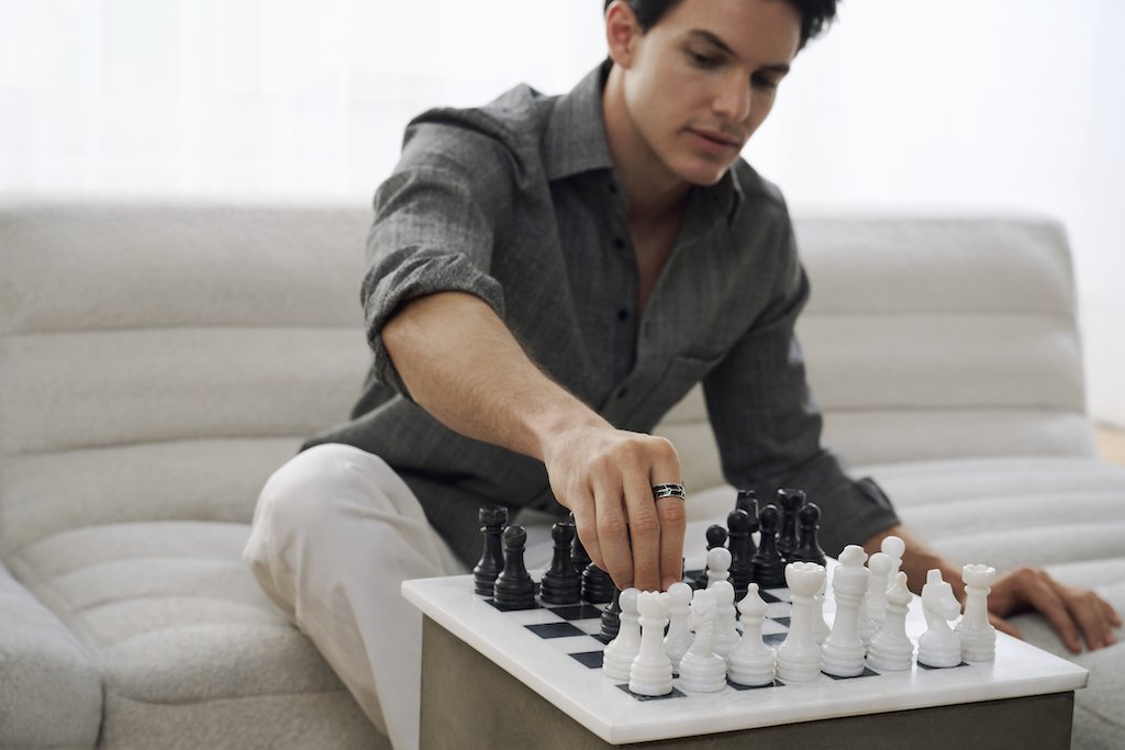 A man wearing a black and silver ring in a gray button-down shirt and white pants sits on a light beige sofa, reaching for a black chess piece on a chessboard with white pawns.