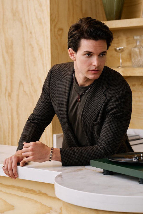 A man wearing a wooden and gold bracelet, a golden pinky ring and a wooden tag on a leather necklace along with a textured black blazer over a dark shirt leans on a white counter in a modern room with light wood paneling. A green turntable is on the counter next to him.
