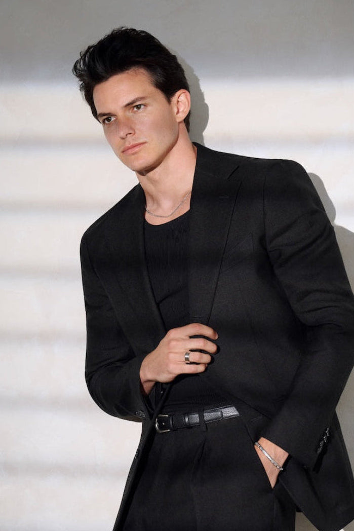 A man wearing a silver band ring, silver box chain and silver bracelet along with a black blazer over a black crew-neck t-shirt and matching black trousers, standing against a light gray wall with soft horizontal shadows.