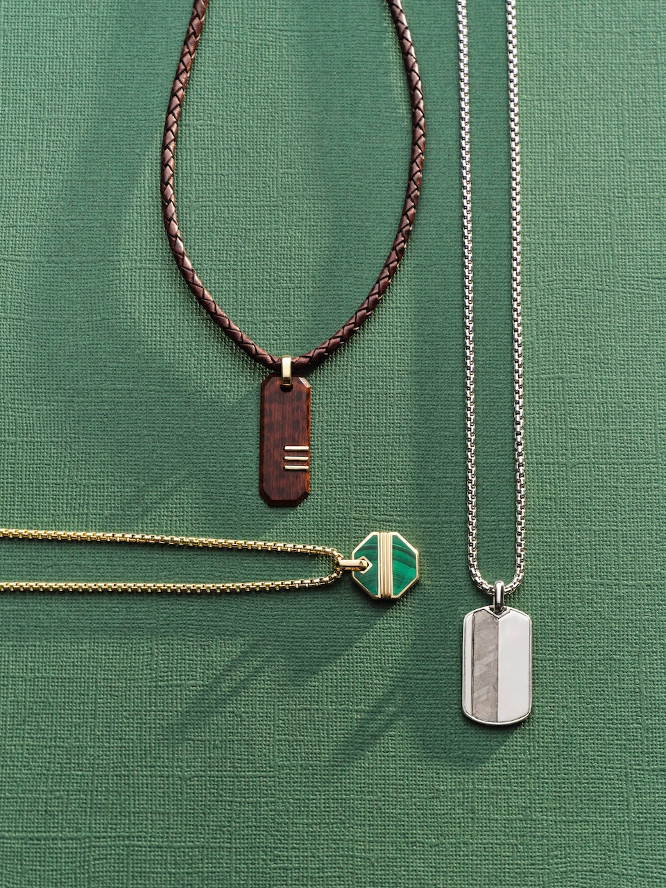 Three necklaces with different pendants on a green textured background: a silver pendant with a meteorite inlay, a gold pendant with a malachite inlay and a wood pendant with gold inlays