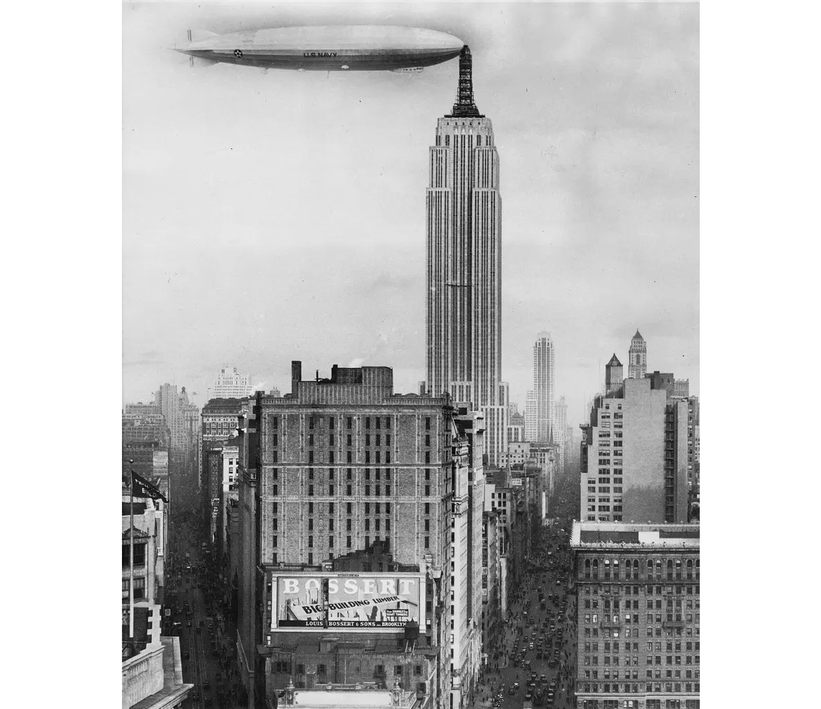 Black and white historical photograph of the New York City skyline, featuring the Empire State Building and a large airship docked at its spire.