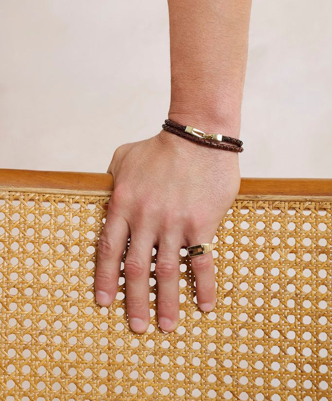 A close-up of a man's hand resting on a woven cane clutch, wearing a dark brown leather bracelet with gold hardware and a matching gold signet ring with tiger's eye inlay.