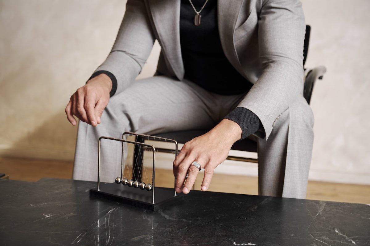 A man wearing a silver ring and silver tag with a light gray suit sits at a table and touches a Newton's cradle with metal balls. The background is a softly lit beige wall.